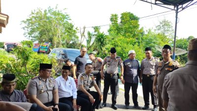 Sekelompok pria, sebagian besar berseragam Polisi RI dan satu berseragam Satpol PP, duduk dan berdiri bersama di area luar ruangan yang teduh di depan sebuah rumah. Seorang pria berbaju koko abu-abu dan peci hitam duduk di tengah, tampaknya sedang bercakap-cakap. Ada papan bunga dukacita di latar belakang.