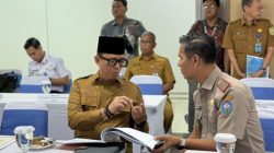 Bupati Ogan Komering Ulu Teddy Meilwansyah yang mengenakan seragam PNS dan peci hitam, sedang berdiskusi serius sambil menunjuk dokumen dengan seorang pejabat militer di meja rapat formal yang memiliki papan nama "BUPATI OGAN KOMERING ULU".