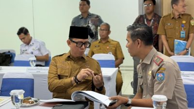 Bupati Ogan Komering Ulu Teddy Meilwansyah yang mengenakan seragam PNS dan peci hitam, sedang berdiskusi serius sambil menunjuk dokumen dengan seorang pejabat militer di meja rapat formal yang memiliki papan nama "BUPATI OGAN KOMERING ULU".