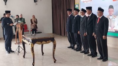 Bupati OKU Teddy Meilwansyah sedang membacakan naskah pelantikan di depan lima pengurus baru Baznas OKU yang mengenakan jas hitam dan peci.