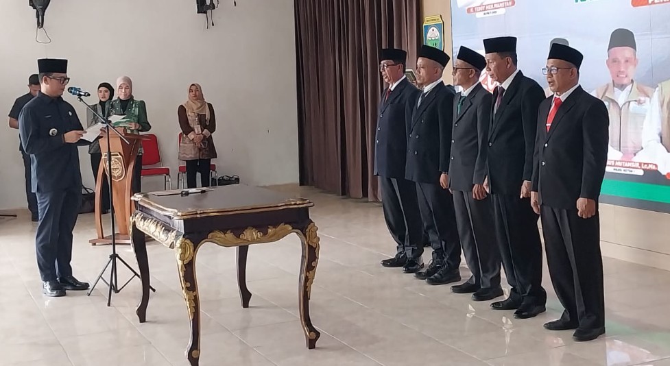 Bupati OKU Teddy Meilwansyah sedang membacakan naskah pelantikan di depan lima pengurus baru Baznas OKU yang mengenakan jas hitam dan peci.