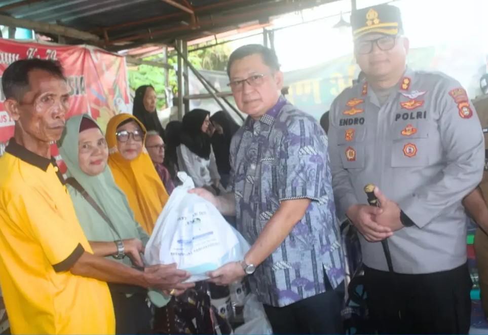 Foto Wakil Bupati OKU (batik ungu) memberikan kantong plastik bantuan putih kepada pria tua (baju kuning), di sebelah kanan berdiri Kapolres OKU berseragam polisi, di depan spanduk merah perayaan Dirgahayu RI.