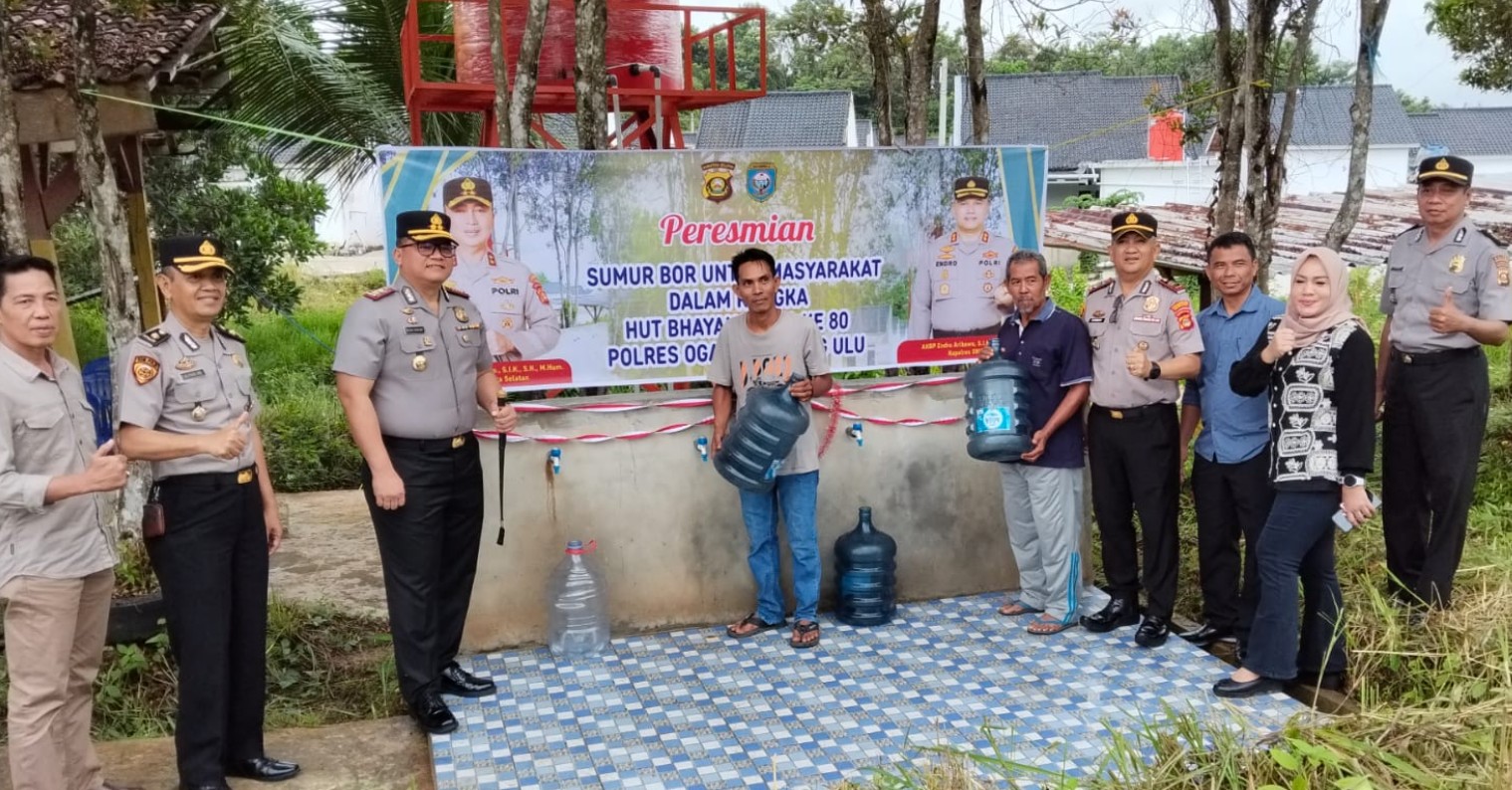  Sekelompok polisi dan warga berpose di depan spanduk peresmian sumur bor. Di belakang mereka terdapat menara tangki air merah dan warga memegang galon air kosong.