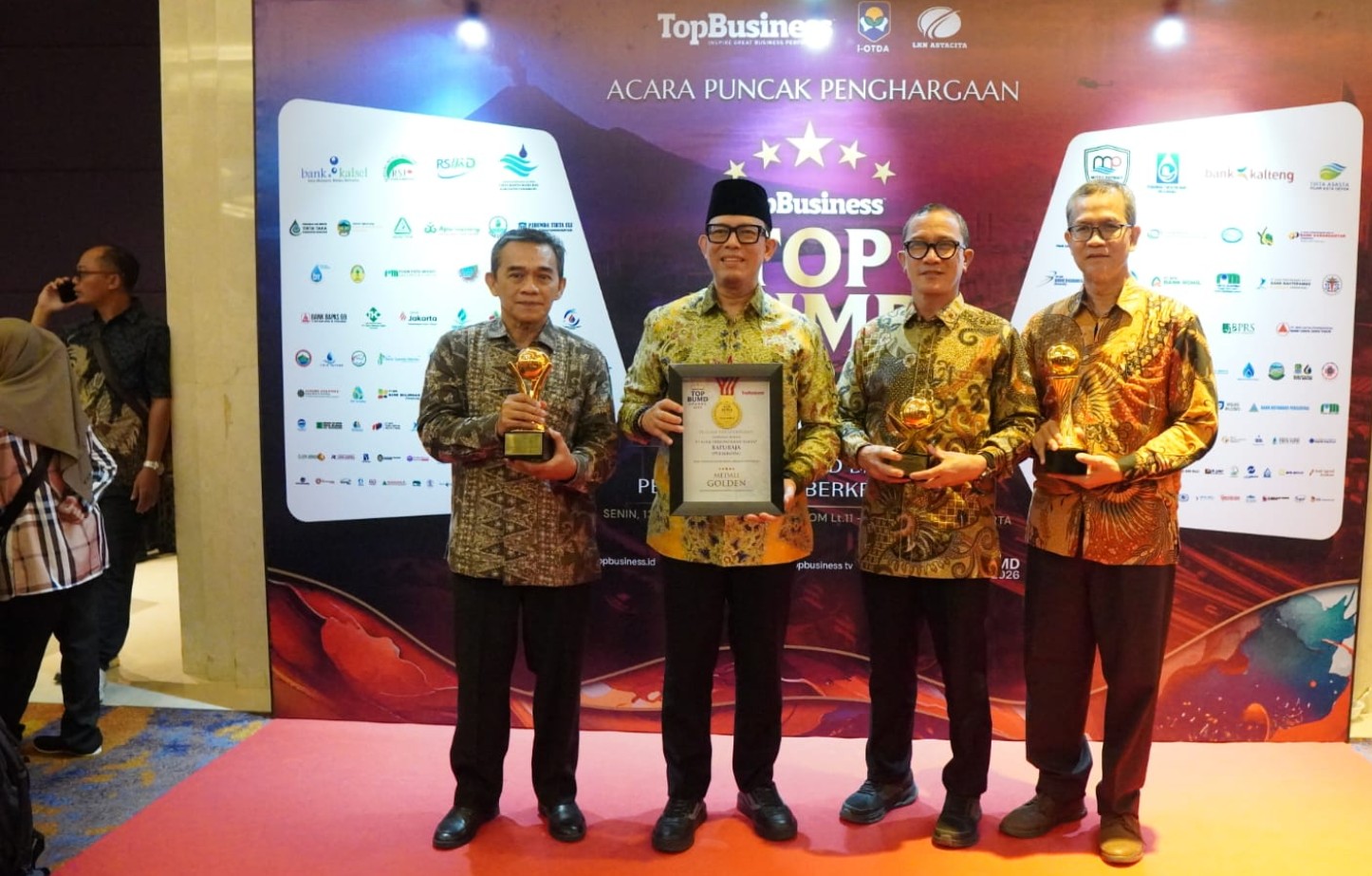 Empat pria mengenakan kemeja batik berdiri di depan backdrop acara 'TOP BUMD Awards'. Pria di tengah berpeci memegang piagam, pria lainnya memegang trofi emas.