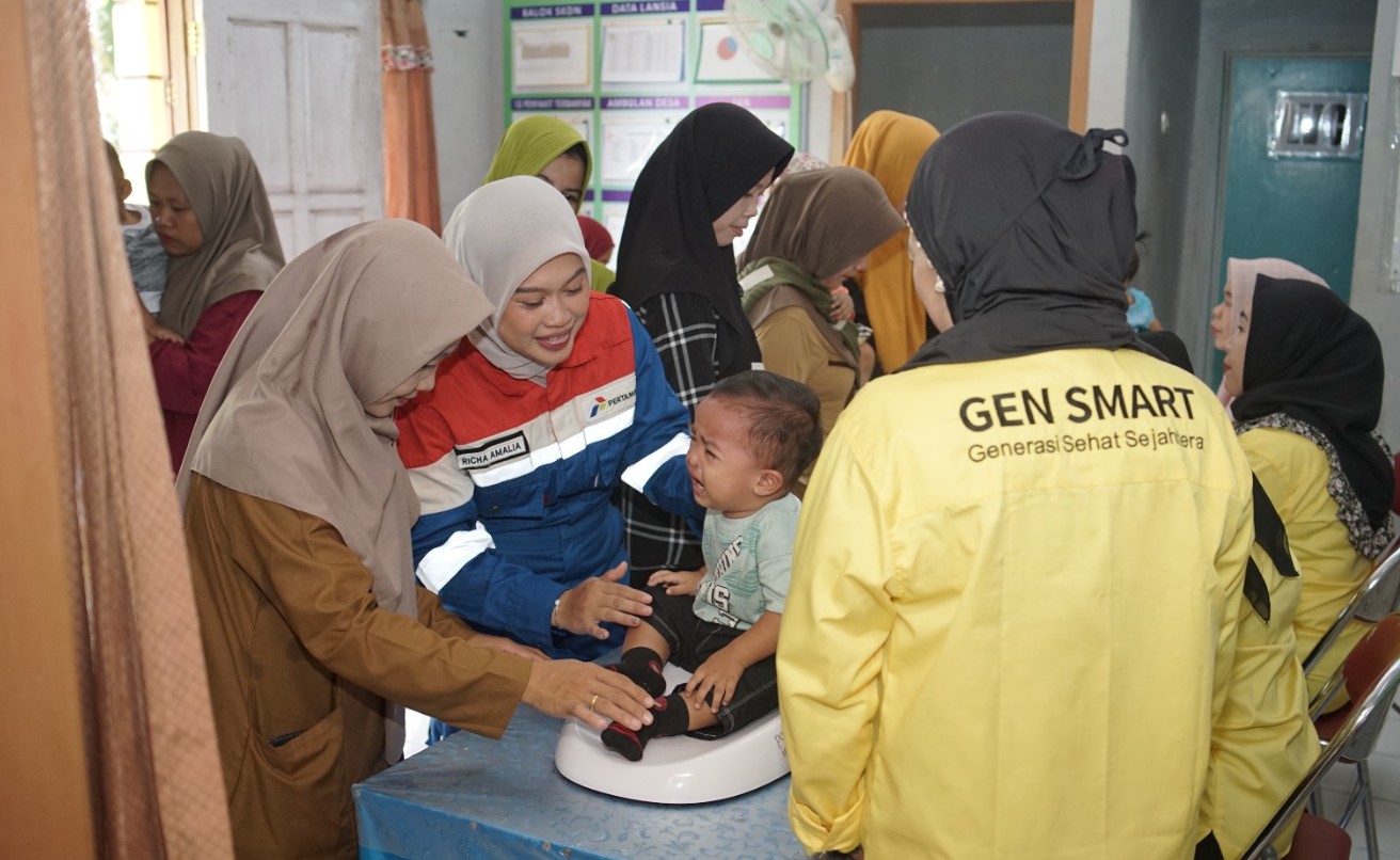 Seorang staf Pertamina berhijab menimbang seorang balita yang menangis di timbangan bayi, sementara seorang petugas kesehatan berjaket kuning bertuliskan "GEN SMART" membantu.