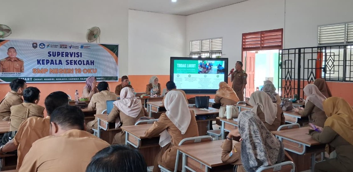 Foto suasana ruang kelas supervisi dengan puluhan guru seragam cokelat duduk di meja, seorang pengawas pria memaparkan materi di depan layar besar, dan spanduk supervisi di latar belakang.