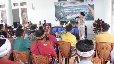 Bupati Sumba Barat Daya, Ratu Ngadu Bonnu Wulla menyampaikan kepada warga latar belakang bertuliskan 'SOSIALISASI PENANGANAN LONG SEGMEN JALAN OMBARADE - KALIMBU TILLU'. Audiens difoto dari belakang, beberapa mengenakan penutup kepala tradisional Sumba. Latar belakangnya adalah bangunan semi-terbuka dengan tanaman hias.