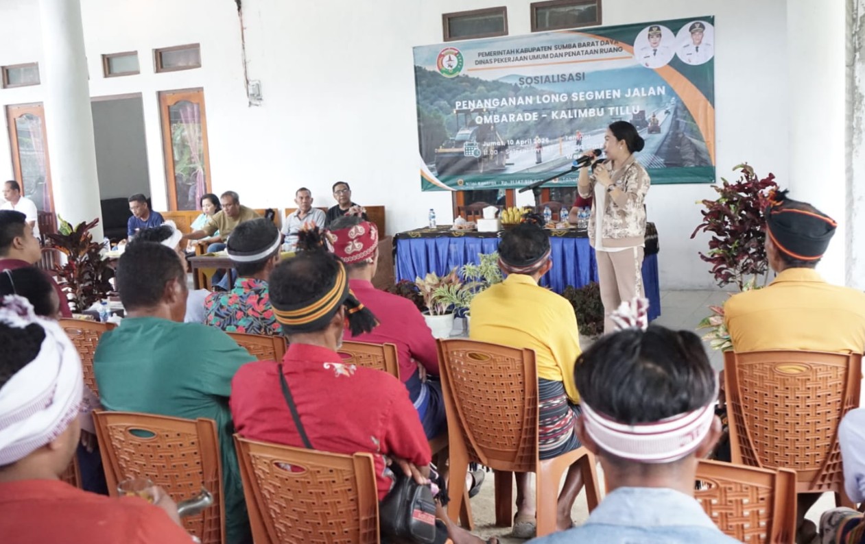 Bupati Sumba Barat Daya, Ratu Ngadu Bonnu Wulla menyampaikan kepada warga latar belakang bertuliskan 'SOSIALISASI PENANGANAN LONG SEGMEN JALAN OMBARADE - KALIMBU TILLU'. Audiens difoto dari belakang, beberapa mengenakan penutup kepala tradisional Sumba. Latar belakangnya adalah bangunan semi-terbuka dengan tanaman hias.