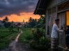 Pemandangan senja di pedesaan Indonesia saat hujan. Seorang kakek bersarung dan kopiah bersama cucu perempuannya berdiri di pintu rumah panggung kayu yang terang oleh lampu teplok. Di latar belakang, terlihat sawah hijau, pohon kelapa, dan sebuah masjid dengan menara yang menyala di bawah langit jingga mendung.