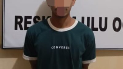 Pria berkaus hijau gelap dengan tulisan 'CONVERSE' dan kerah putih berdiri tegak dengan tangan di belakang, wajahnya disensor. Ia berada di depan spanduk berlogo kepolisian bertuliskan 'SIDIK SAKTI INDERA WASPADA' dan sebagian teks 'RESKRIM ULU OGAN'.