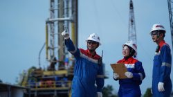 Tiga pekerja Pertamina EP Zona 4 mengenakan alat pelindung diri lengkap sedang berdiskusi dengan latar belakang rig pengeboran sumur migas.