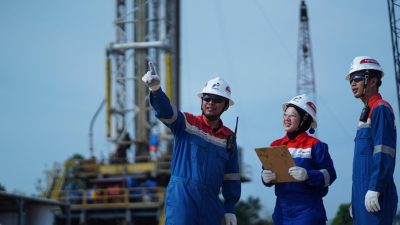 Tiga pekerja Pertamina EP Zona 4 mengenakan alat pelindung diri lengkap sedang berdiskusi dengan latar belakang rig pengeboran sumur migas.