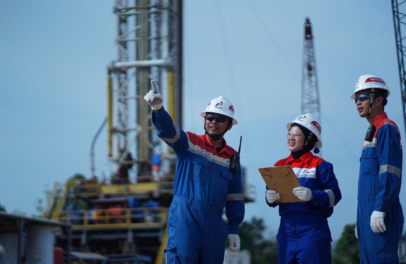 Tiga pekerja Pertamina EP Zona 4 mengenakan alat pelindung diri lengkap sedang berdiskusi dengan latar belakang rig pengeboran sumur migas.