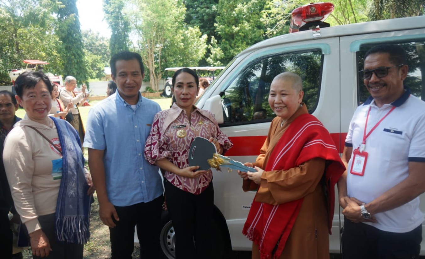 oto close-up lima orang di samping ambulans putih dengan lampu darurat merah. Di tengah, seorang wanita berbatik merah muda (Bupati Ratu Wulla) dan seorang biksu Budha (Dr. Liang) bersama-sama memegang plakat kunci ambulans besar berwarna putih. Mereka tersenyum. Pria berkemeja biru (Wakil Bupati Dominikus) berdiri di dekat mereka.
