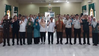 Sekelompok besar orang di aula, dengan 12 pejabat di depan, semuanya mengangkat tangan kanan dengan telapak terbuka dalam pose kampanye. Latar belakang menunjukkan ruang pertemuan dengan logo pemerintah.