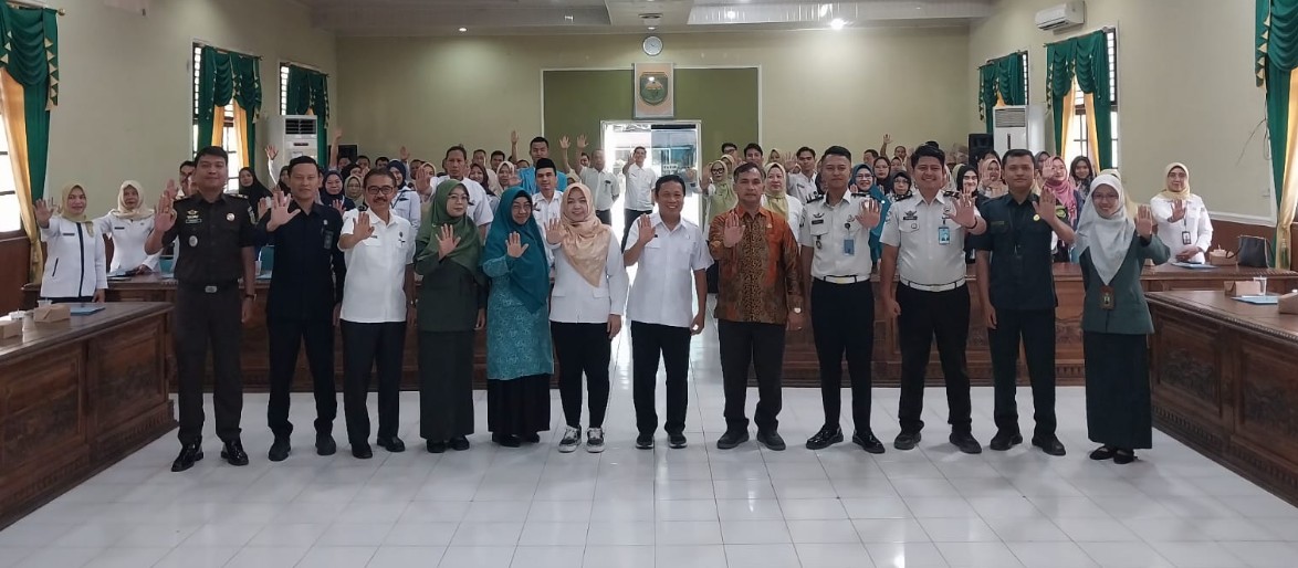 Sekelompok besar orang di aula, dengan 12 pejabat di depan, semuanya mengangkat tangan kanan dengan telapak terbuka dalam pose kampanye. Latar belakang menunjukkan ruang pertemuan dengan logo pemerintah.

