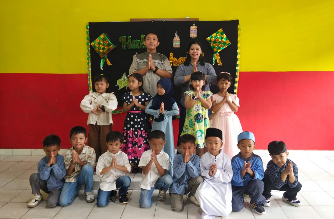Sekelompok siswa dan dua guru berpose salam namaste di depan dekorasi panggung Halal Bihalal.
