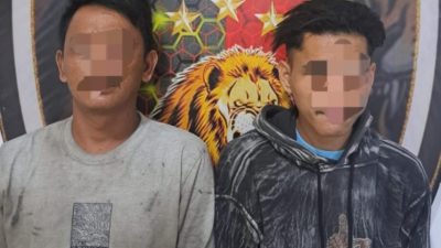 Dua pria muda dengan wajah disensor berdiri berdampingan. Pria di kiri mengenakan kaus abu-abu kotor dengan logo kecil; pria di kanan mengenakan hoodie bermotif hitam-putih dengan grafik tangan di bagian depan. Mereka berdiri di depan spanduk latar belakang pers kepolisian yang menampilkan logo singa.