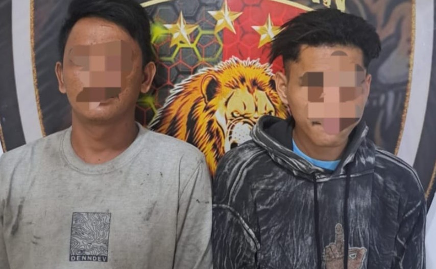 Dua pria muda dengan wajah disensor berdiri berdampingan. Pria di kiri mengenakan kaus abu-abu kotor dengan logo kecil; pria di kanan mengenakan hoodie bermotif hitam-putih dengan grafik tangan di bagian depan. Mereka berdiri di depan spanduk latar belakang pers kepolisian yang menampilkan logo singa.