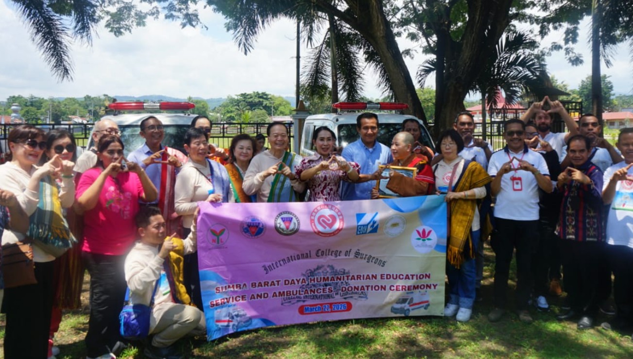 Foto grup besar orang-orang yang berdiri di halaman dengan pepohonan. Mereka memegang spanduk panjang berwarna-warni yang mengumumkan upacara donasi ambulans untuk Sumba Barat Daya. Di spanduk terdapat teks "International College of Surgeons, SUMBA BARAT DAYA HUMANITARIAN EDUCATION SERVICE AND AMBULANCES DONATION CEREMONY, March 27, 2026". Beberapa orang membuat tanda hati dengan tangan. Dua ambulans putih dengan lampu darurat terlihat di latar belakang.
