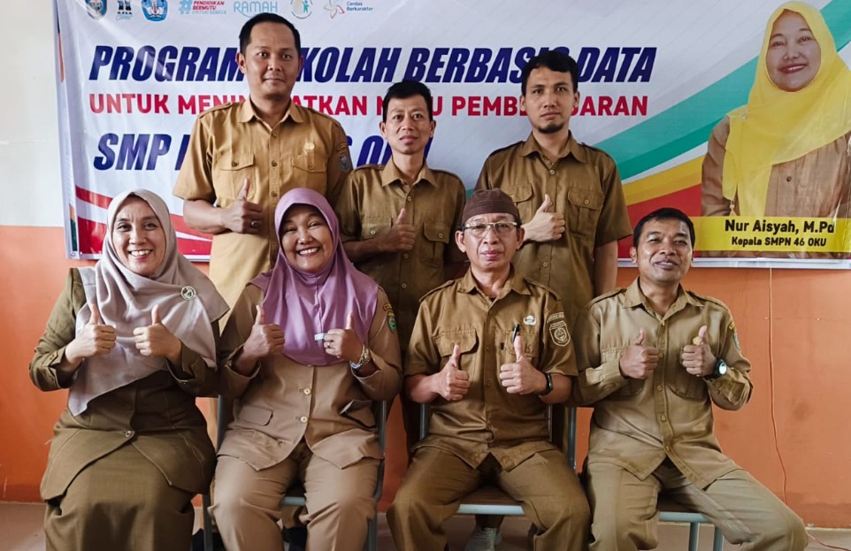 Sekelompok orang berseragam dinas guru memberikan acungan jempol dengan spanduk "Program Sekolah Berbasis Data SMP Negeri 46 OKU" di latar belakang.
