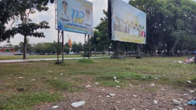 A photo of an open park area in Indonesia with two large metal-framed billboards in the background, featuring photos of officials and text commemorating Indonesian events, with plastic trash scattered across the grass in the foreground under a cloudy sky, dated June 29, 2026.