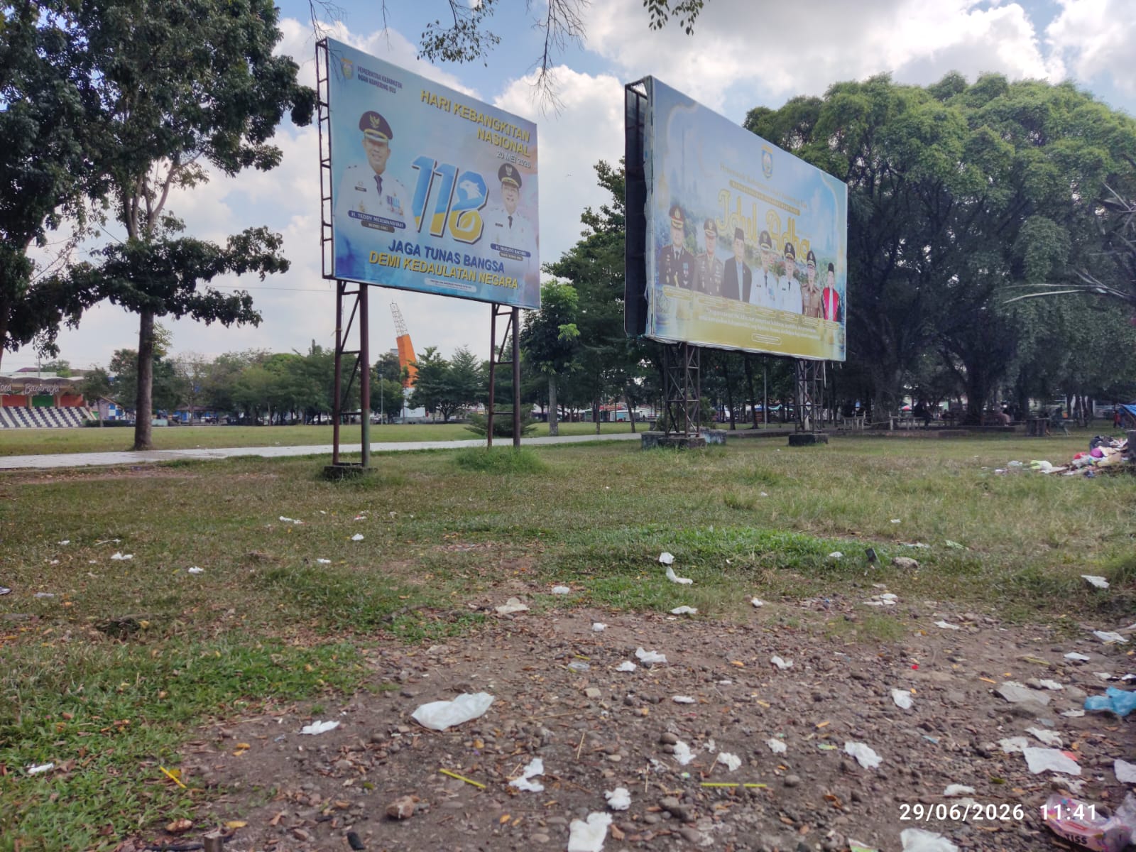 A photo of an open park area in Indonesia with two large metal-framed billboards in the background, featuring photos of officials and text commemorating Indonesian events, with plastic trash scattered across the grass in the foreground under a cloudy sky, dated June 29, 2026.