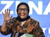 Potret Nanik S Deyang tersenyum dan melambaikan tangan mengenakan jilbab hitam dan kemeja batik, berlatar belakang biru.