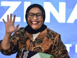 Potret Nanik S Deyang tersenyum dan melambaikan tangan mengenakan jilbab hitam dan kemeja batik, berlatar belakang biru.