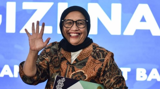 Potret Nanik S Deyang tersenyum dan melambaikan tangan mengenakan jilbab hitam dan kemeja batik, berlatar belakang biru.