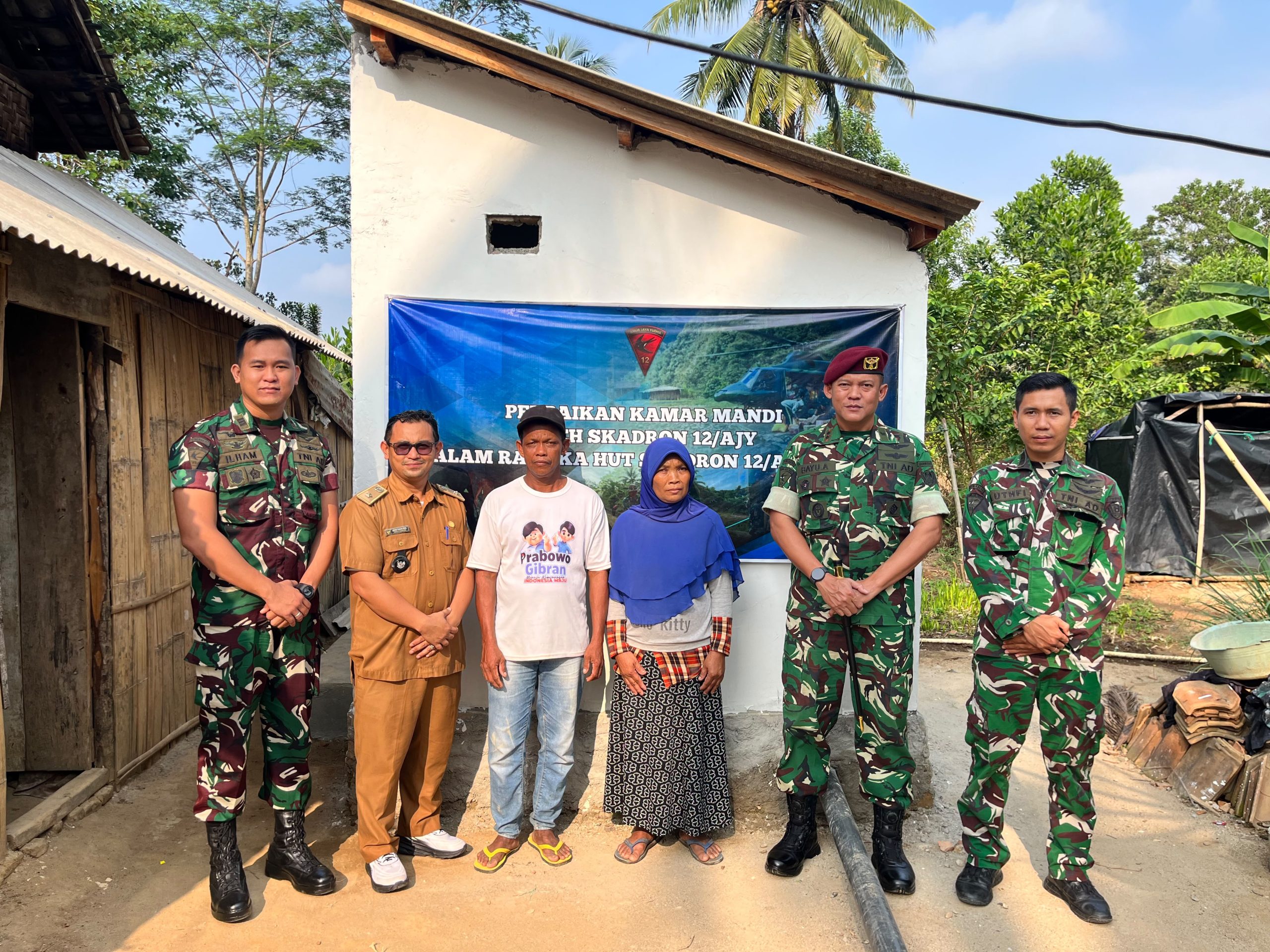 ultah 14 Skadron 12 Amur Jaya Yudha.jpg, adalah foto bersama yang lebih besar yang diambil di depan teras sebuah bangunan (posko atau bangunan fasilitas umum). Sekelompok personel TNI, baik yang berdiri maupun jongkok, bersama beberapa warga sipil, berpose serempak dengan simbol tangan "persatuan" (tangan disilangkan di dada). Di belakang mereka, spanduk besar dan jelas bertuliskan "KARYA BAKTI TNI DALAM RANGKA HUT KE-14 SKADRON 12/AMU JAYA YUDHA". Foto ini menunjukkan semangat kebersamaan dan kekompakan antara TNI dan masyarakat. Bangunan di belakang memiliki atap genteng merah yang khas.