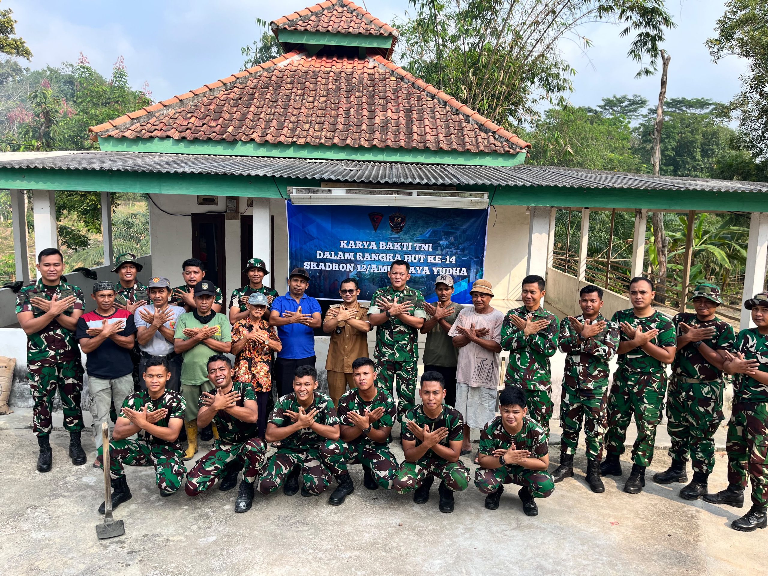 Empat personel TNI berseragam, termasuk seorang Letkol berseragam Baret Merah, berdiri bersama seorang pejabat pemerintah setempat (baju dinas cokelat) dan dua warga penerima manfaat (pria dengan kemeja kampanye dan wanita berhijab). Mereka berpose di depan spanduk yang menyebutkan "Perbaikan Kamar Mandi Skadron 12/AJY" dalam rangka ulang tahun mereka, di samping bangunan kamar mandi yang terlihat baru dicat putih. Latar belakangnya menunjukkan lingkungan pedesaan yang asri dengan pohon bambu.