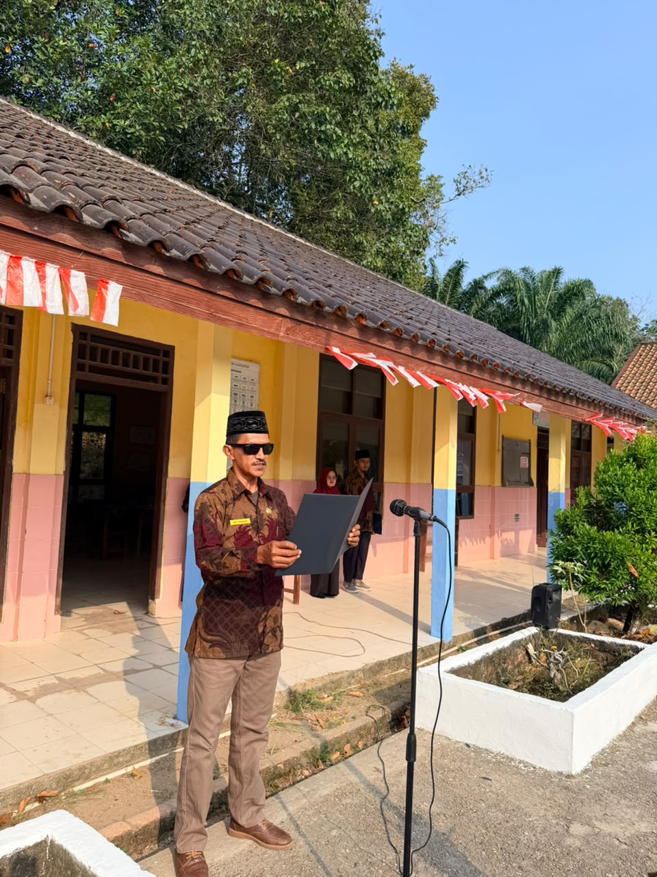 Seorang pria mengenakan peci hitam, kacamata hitam, dan seragam batik lengan panjang sedang membacakan teks dari map biru di depan mikrofon di luar ruangan.
