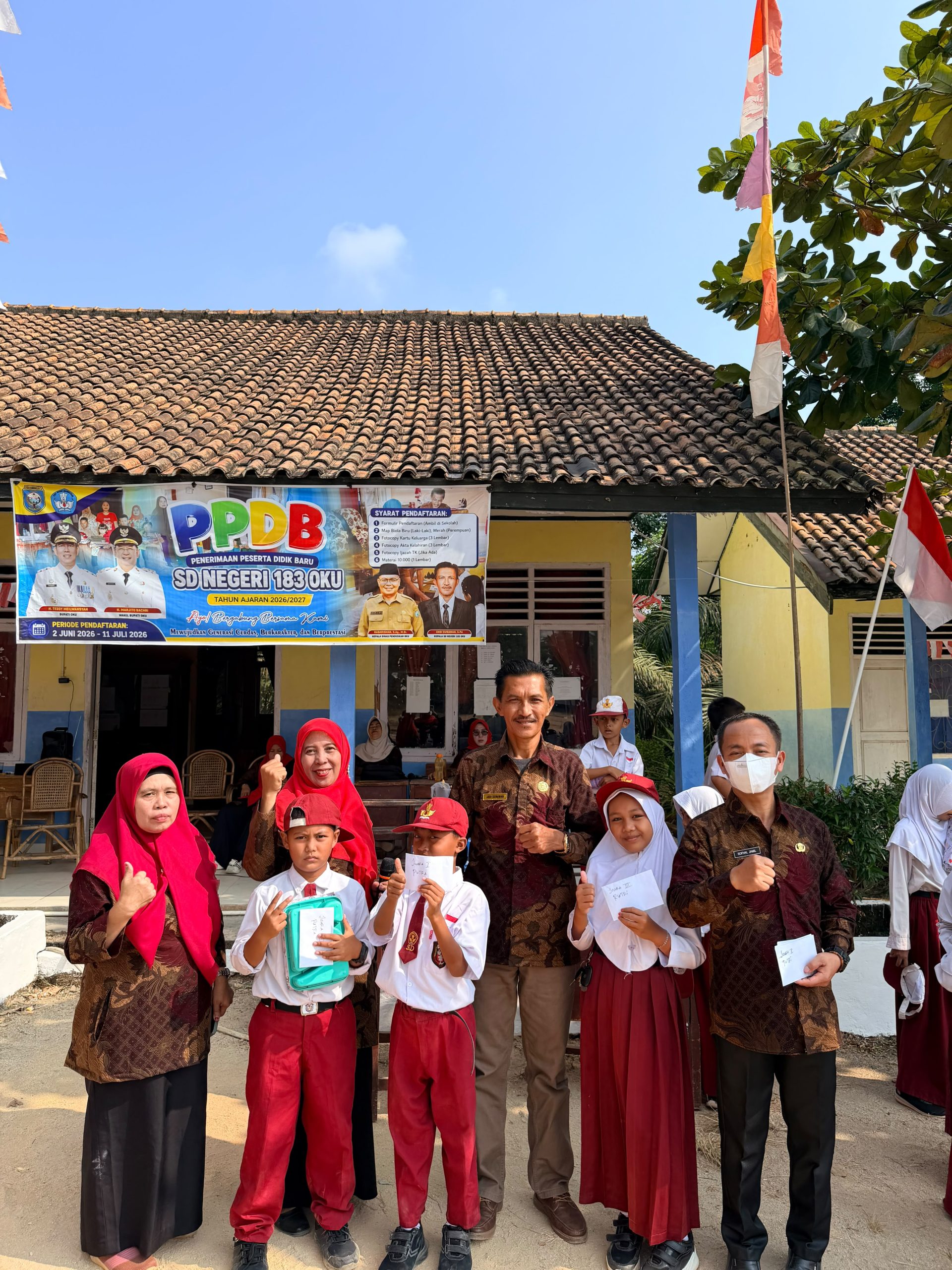Sekelompok guru dan siswa SD Negeri 183 OKU berpose ceria di depan gedung sekolah yang dihiasi spanduk PPDB dan bendera Merah Putih.