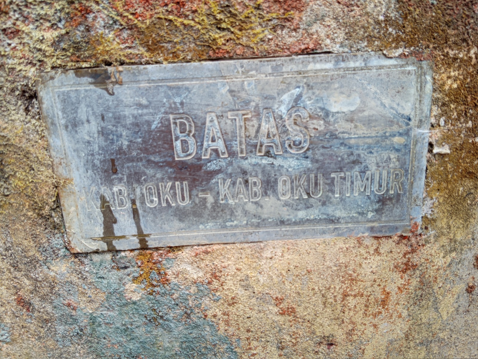 Foto memotret secara dekat sebuah pelat logam berbentuk persegi panjang yang ditempelkan pada struktur beton atau batu berserat kasar dan agak berlumut. Pada plakat tersebut tercetak huruf timbul yang berbunyi "BATAS" di baris pertama, dan "KAB. OKU - KAB. OKU TIMUR" di baris kedua.