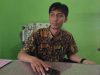 Kabid Administrasi Pemerintahan Desa PMD OKU, M Rizal Ramadhoni, tersenyum mengenakan kemeja batik saat duduk di meja kerjanya dengan latar belakang dinding hijau.