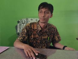 Kabid Administrasi Pemerintahan Desa PMD OKU, M Rizal Ramadhoni, tersenyum mengenakan kemeja batik saat duduk di meja kerjanya dengan latar belakang dinding hijau.