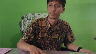 Kabid Administrasi Pemerintahan Desa PMD OKU, M Rizal Ramadhoni, tersenyum mengenakan kemeja batik saat duduk di meja kerjanya dengan latar belakang dinding hijau.