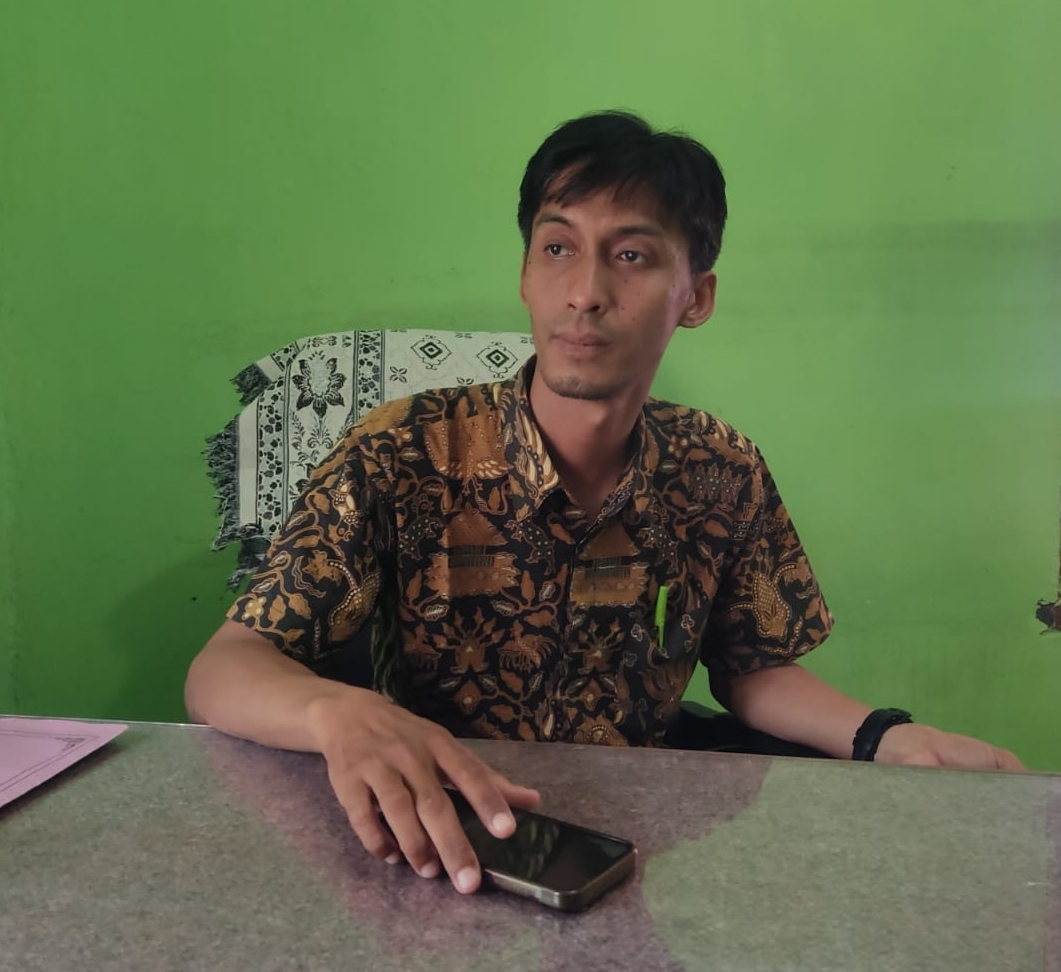 Kabid Administrasi Pemerintahan Desa PMD OKU, M Rizal Ramadhoni, tersenyum mengenakan kemeja batik saat duduk di meja kerjanya dengan latar belakang dinding hijau.