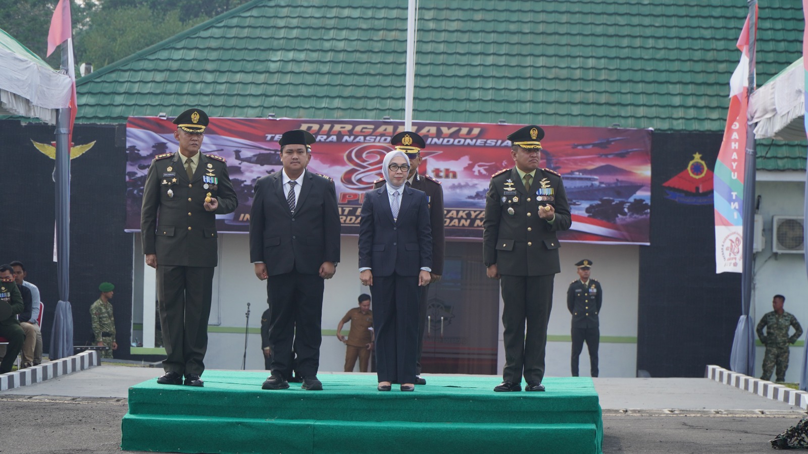 Pejabat militer dan pejabat sipil berdiri tegap di atas mimbar podium hijau pada upacara resmi.