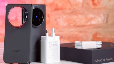 Ponsel pintar lipat Vivo X Fold 6 berwarna hitam dalam posisi berdiri di samping adaptor pengisi daya berwarna putih, gulungan kabel data putih, dan kotak kemasan hitam bertuliskan vivo X Fold6.