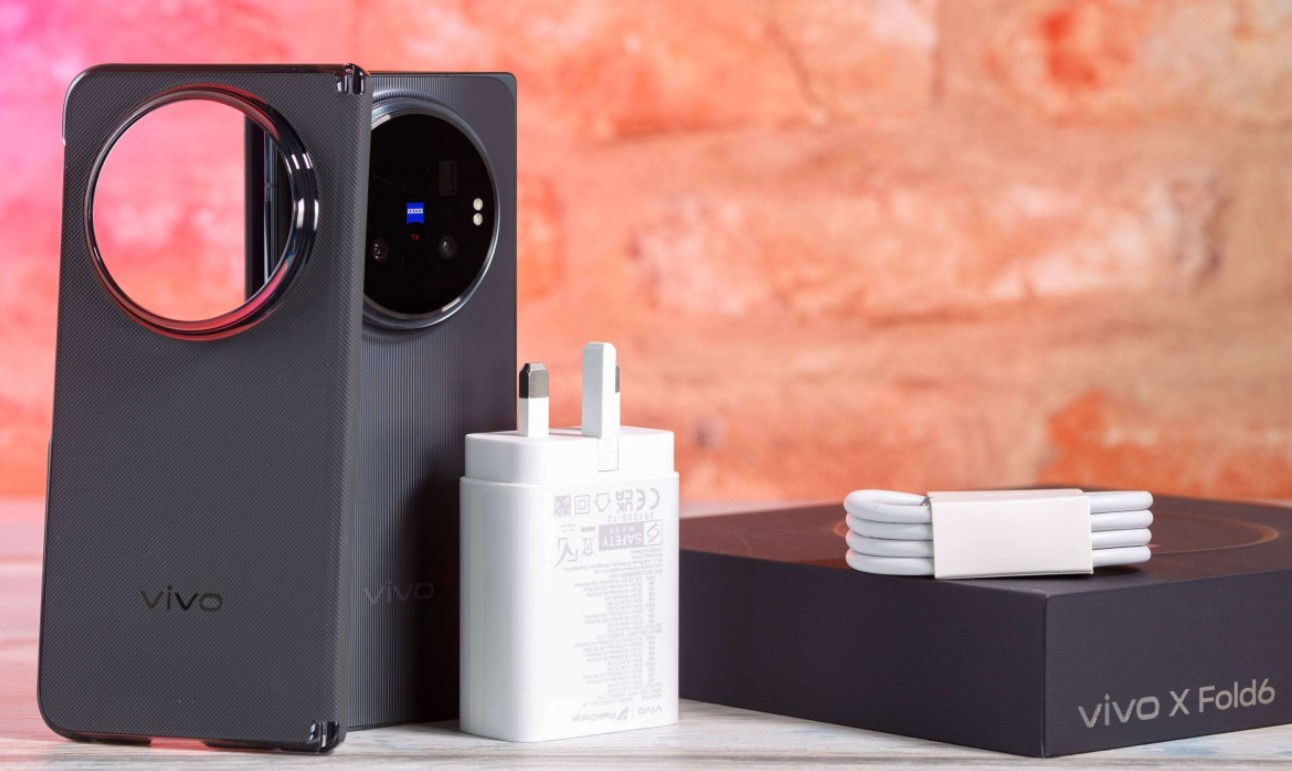 Ponsel pintar lipat Vivo X Fold 6 berwarna hitam dalam posisi berdiri di samping adaptor pengisi daya berwarna putih, gulungan kabel data putih, dan kotak kemasan hitam bertuliskan vivo X Fold6.