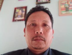 Potret seorang pria paruh baya berkumis yang mengenakan jas hitam dan kemeja bergaris gelap, berfoto dengan latar belakang dinding berwarna biru muda yang dihiasi beberapa bingkai foto.