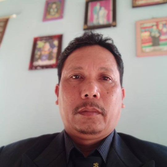 Potret seorang pria paruh baya berkumis yang mengenakan jas hitam dan kemeja bergaris gelap, berfoto dengan latar belakang dinding berwarna biru muda yang dihiasi beberapa bingkai foto.